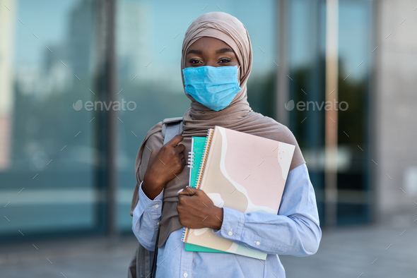Study During Coronavirus. African Muslim Female Student In Medical Mask And Hijab Posing Outdoors With Books, Standing Near University Building, Resting Between Classes, Looking At Camera, Copy Space
