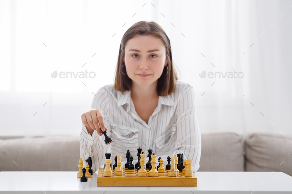 Board games. Portrait of young woman plays in chess and looks at camera on window background at home, copy space