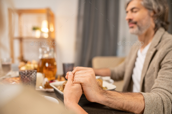 Hands of mature man with closed eyes and his wife praying by served table