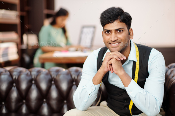 Portrait of smiling handsome young Indian tailor with measuring tape sitting on sofa in atelier and looking at camera