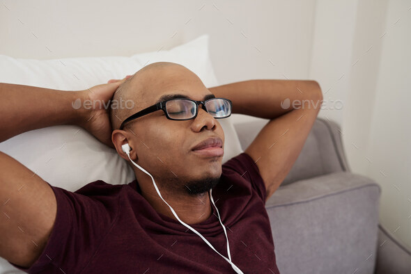 Tired young Black man in glasses closing eyes when relaxing on sofa and enjoying good music in earbuds