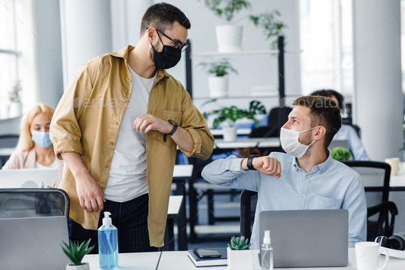 Safe hello at work during COVID-19 epidemic and social distance. Young men in protective masks greet with elbows at workplace with laptop and antiseptic in modern office interior, copy space