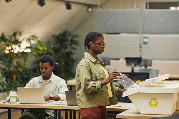 Side view portrait of young African-American woman putting paper cup into waste sorting bin in office, copy space