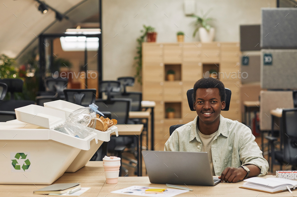 Portrait of smiling African-American man using laptop while sitting by waste sorting bins in modern office, copy space