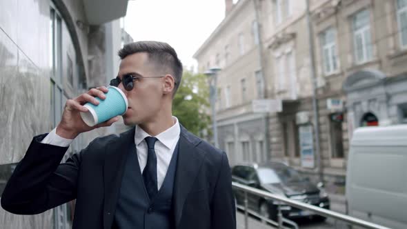 Man in Suit with Sunglasses Drinks a Cup of Beverage When Looks Around on Street alt