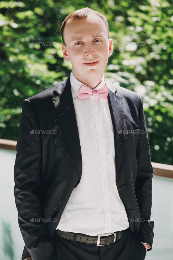 Stylish Groom Putting On Pink Bow Tie And Posing In Black Suit Outdoors  Stock Photo By Sonyachny