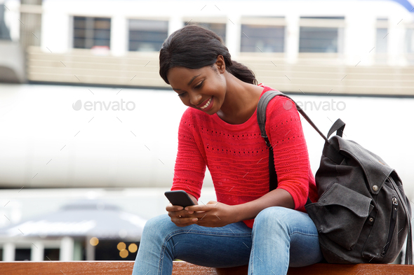 smiling young african american woman sitting outside in the city with bag and cellphone - Stock Photo - Images