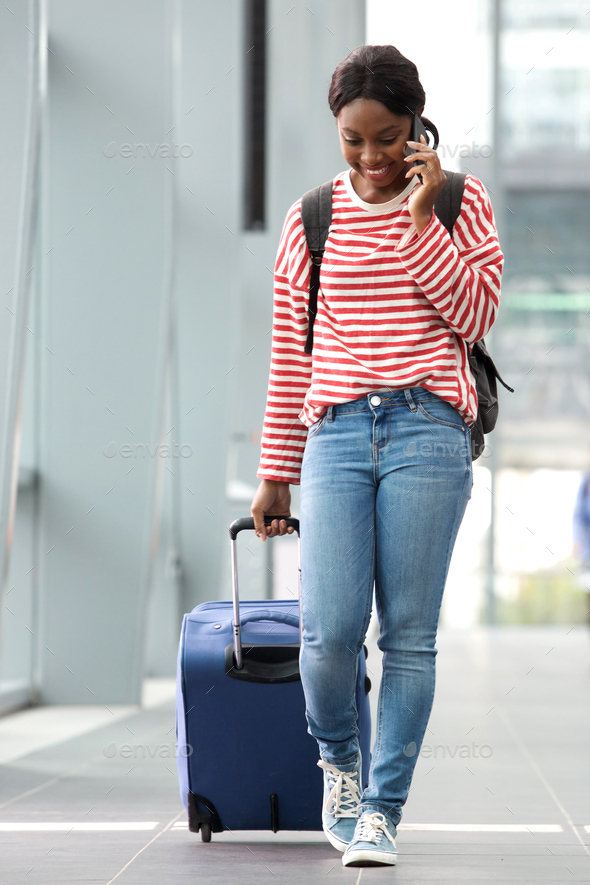 Full length portrait of young female traveler walking in airport terminal with suitcase and mobile phone