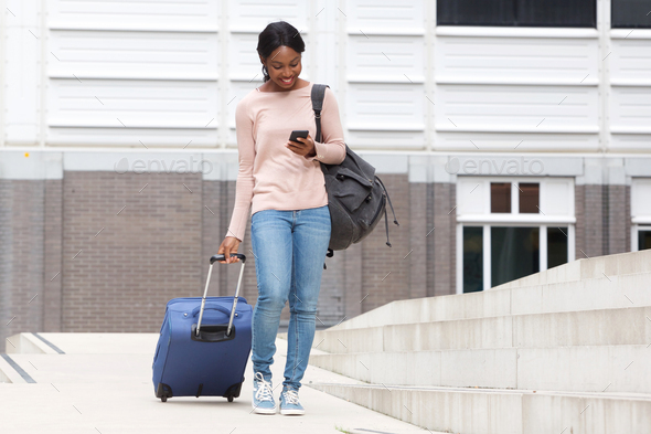 Full body happy young african american woman walking with suitcase bag and cellphone - Stock Photo - Images