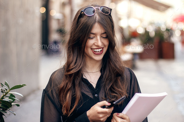 beautiful stylish hipster girl smiling in sunny city street, holding magazine - Stock Photo - Images