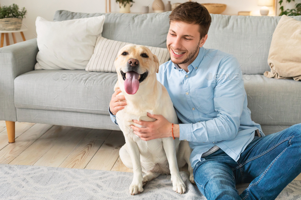 Contact With Animal Concept. Portrait of handsome guy owner hugging his golden retriever sitting on the floor in living room. Happy man spending time indoors at home in modern apartment
