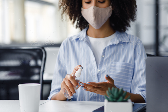 African american woman in protective mask using antiseptic to disinfect hands at workplace in - Stock Photo - Images