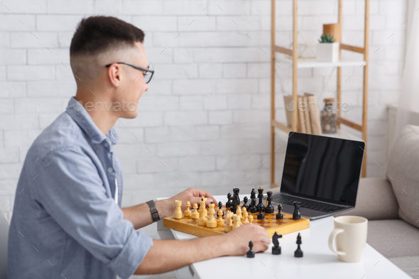 Smart guy and hobby in free time on self-isolation. Happy chess player in glasses with chess board looking at laptop with blank screen in living room interior during coronavirus outbreak, copy space