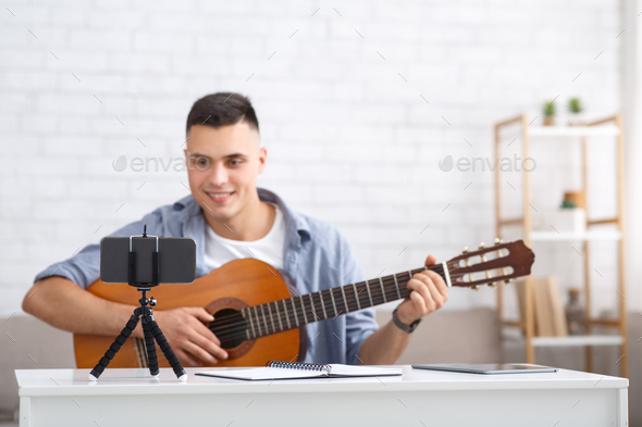 Leisure time, hobby and modern technology. Cheerful millennial man plays on guitar and looks at web - Stock Photo - Images
