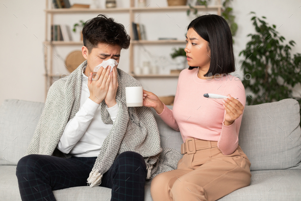 Supportive Asian Wife Treating Sick Husband With Flu Giving Him Medicine Sitting On Couch At Home. Korean Girlfriend Takes Care Of Ill Boyfriend. Seasonal Influenza, Cold, Illness Concept