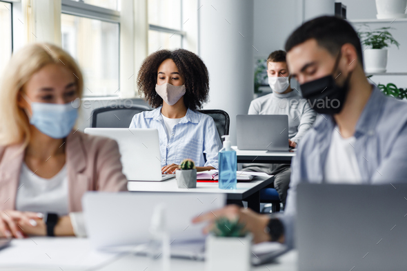 Work with documents and discussion at coronavirus. Guy and woman in protective masks are looking at paper, close up, blurred, focus on african american lady typing on laptop at workplace in office interior