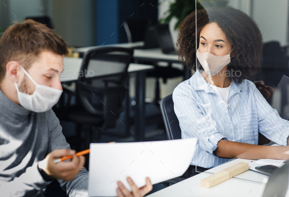 Work with papers and business communication of colleagues. European guy and african american woman in protective masks look at documents at workplace and discus of marketing plan during pandemic covid-19