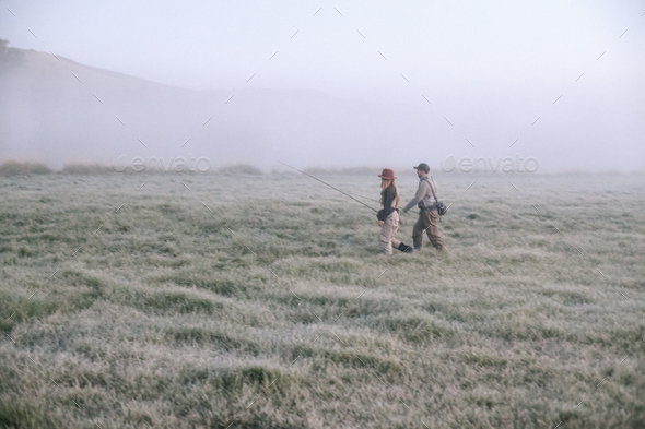 Two people walking across a meadow in early morning mist carrying fishing rods. - Stock Photo - Images
