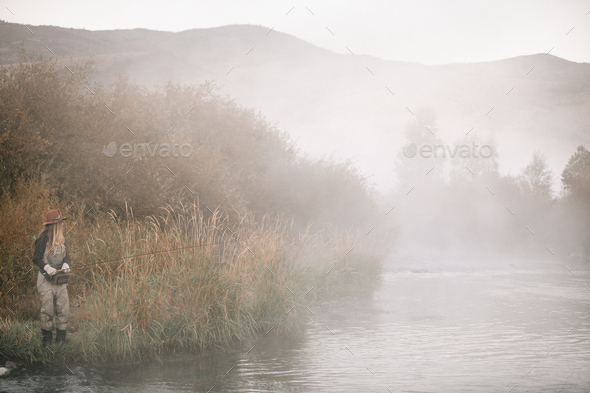 A woman fishing, standing on the riverbank. Mist rising from the water.