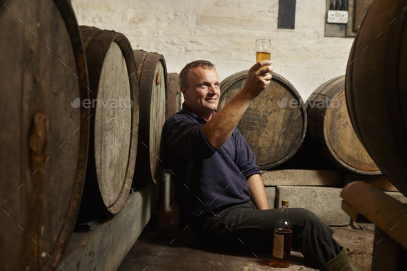 A man sitting among oak barrels at a cider makers, raising a glass and tasting the brew.