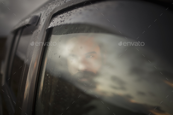 A man sitting in a car looking out. Reflections of the sunset sky on the window.