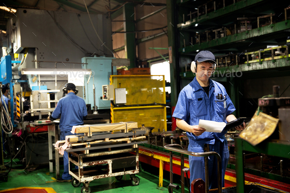 Japanese man standing in factory, holding piece of paper.