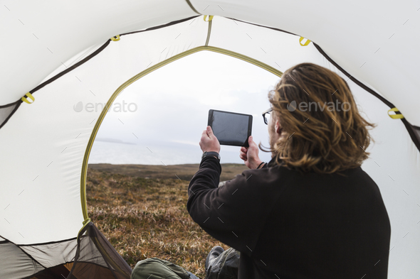 A man sitting in the shelter of a tent looking out, taking a photograph with a digital tablet