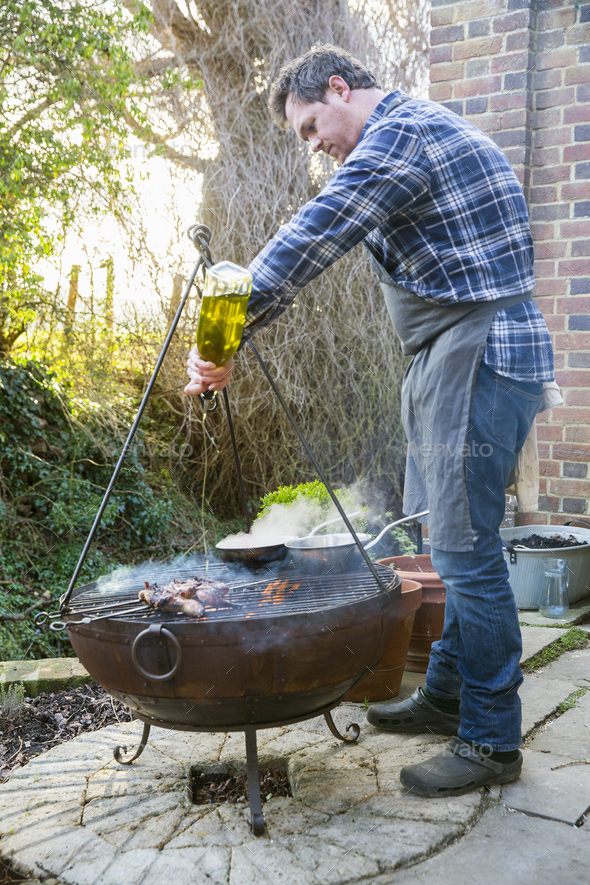 A man cooking roasting game birds over an open fire, pouring olive oil from a bottle on the meat.