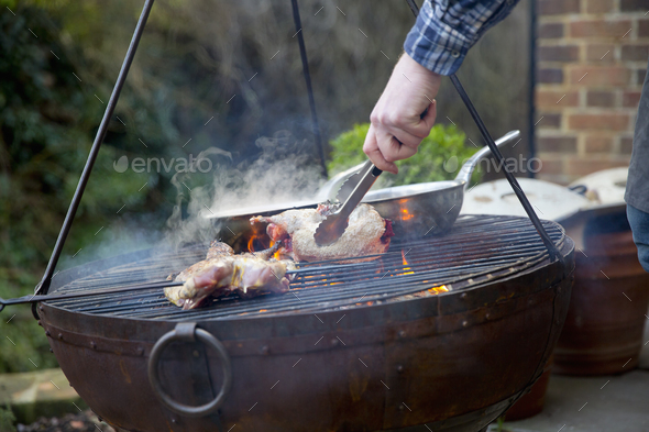 A man using tongs to move roasting game bird pieces on a barbeque.