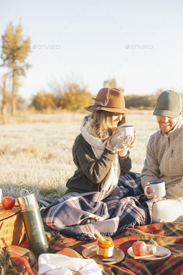 Two people, a couple on a winter picnic, seated on a tartan rug, holding hot drinks.