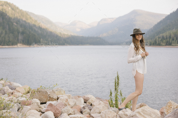 A woman in a hat and white shirt with a basket, by the shore of a mountain lake. - Stock Photo - Images
