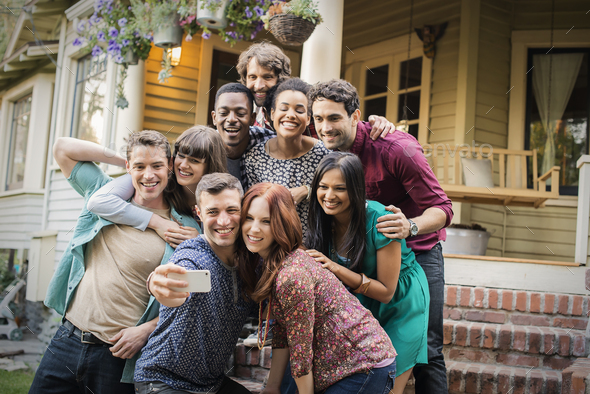 A group of friends posing on the steps of a house porch, taking a group selfy.