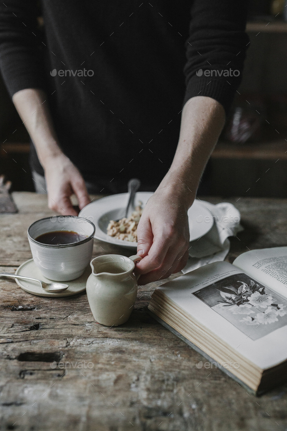 A person at a table with a cup of coffee, bowl of muesli and an open book.