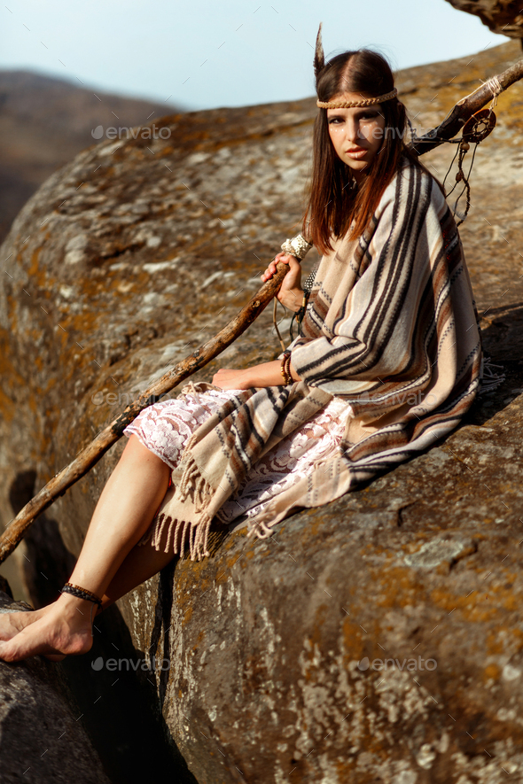 beautiful native indian american woman holding pikestaff sitting on rocks and looking at woods - Stock Photo - Images