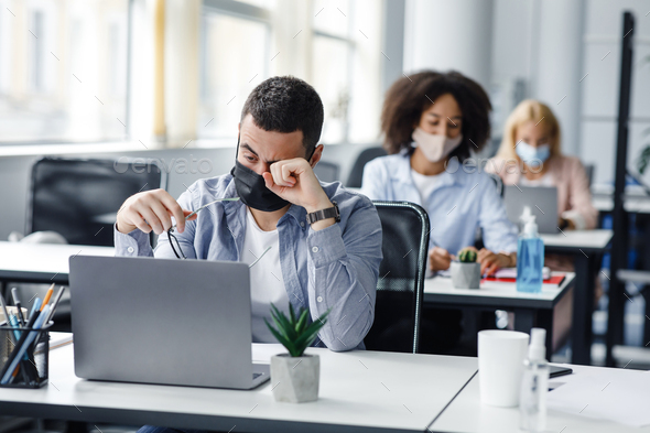 Tired and overworked. Focus on man in protective mask rubs eyes with hand, behind sit female - Stock Photo - Images
