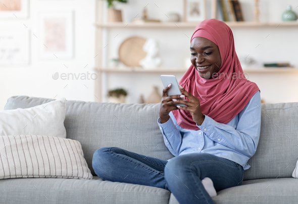 Social Networks. Young black muslim female in hijab using smartphone at home, sitting on cozy couch, messaging with friends, browsing internet, relaxing in living room, enjoying weekend, copy space