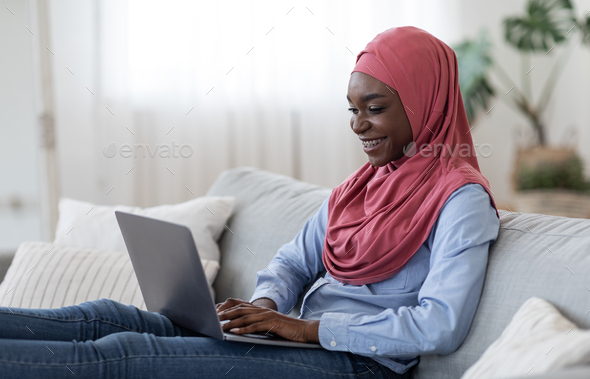 Freelance Concept. Joyful black islamic woman in hijab working remotely on laptop computer at home, typing on keyboard, using modern pc while sitting on comfortable couch in living room, copy space