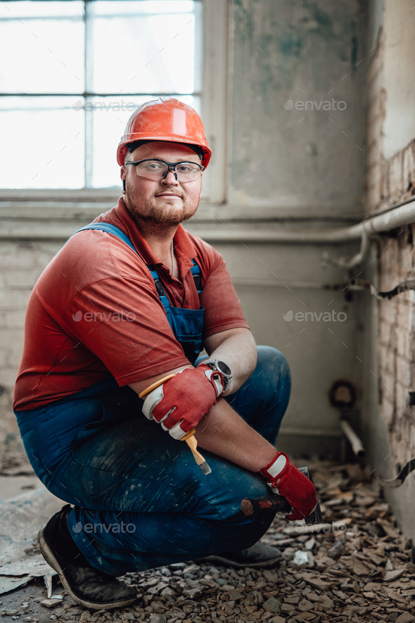 Confident professional worker strikes old plaster from brick wall, holding a hammer