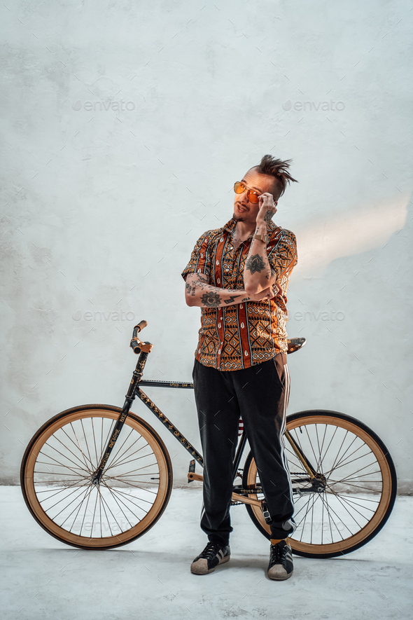 Tattooed gangster freak in hawaiian shirt and sports pants standing next to a custom made bicycle