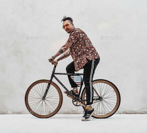 Stylish tattooed guy in Hawaiian shirt and sports pants rides a cool custom made bicycle. Studio shot on a white wall