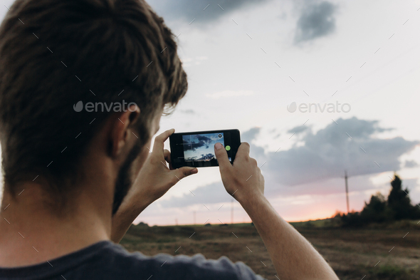 man holding phone and taking photo of amazing sunset landscape view in summer field