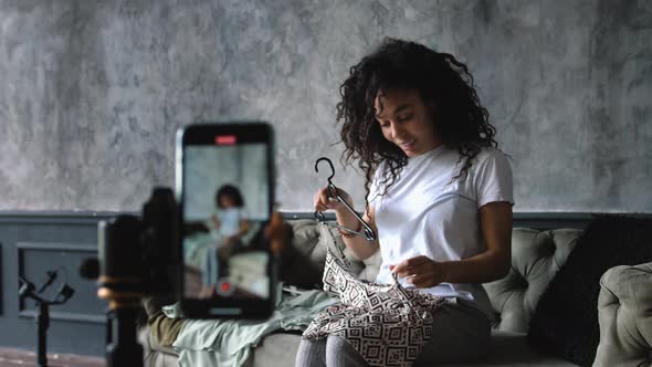 AfricanAmerican Fashion Blogger Recording Video with a Smartphone at Home Choosing Clothes Sitting alt