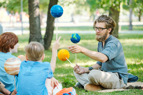 Bearded teacher in eyeglasses pointing at planet model and telling about astronomy to kids at outdoor lesson