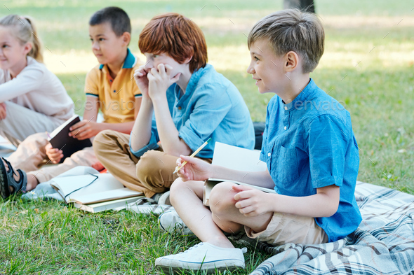 Curious kids sitting with workbooks on plaids while listening to teacher at outdoor lesson