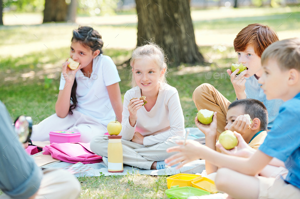 Group of positive kids sitting on plaid and eating apples while listening to teacher at outdoor lesson