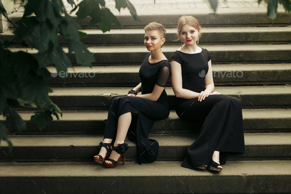 two luxury gorgeous woman in black dress posing, sitting on old stairs in soft light in city, classic gothic lady outfit