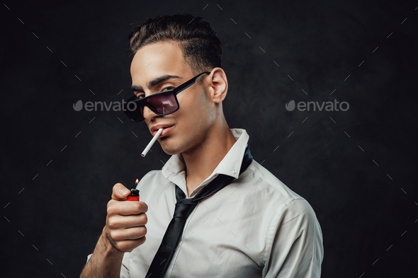 Handsome and successful. Young handsome man in sunglasses wearing white shirt and black tie lights up a cigarette and looking on the camera against black background