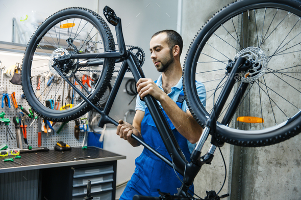 Bicycle repair in workshop, man checks mechanisms. Mechanic in uniform fix problems with cycle, professional bike repairing service
