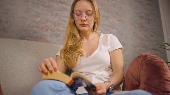 Low Angle Serious Woman Reads Book and Turns Page Sitting on Sofa alt