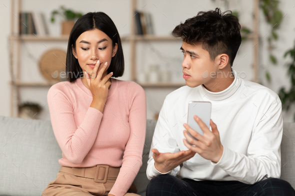 Infidelity In Relationship. Jealous Chinese Husband Showing Cheating Wife Her Cellphone Demanding Explanation Sitting On Couch At Home. Boyfriend Suspecting Unfaithful Girlfriend Having Affair